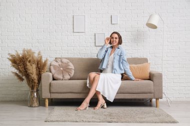 Evdeki beyaz tuğla duvarın yanında gri koltukta oturan genç kadın.