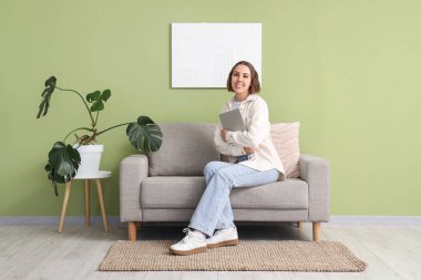 Evde yeşil duvarın yanındaki gri koltukta oturan dizüstü bilgisayarı olan genç bir kadın.
