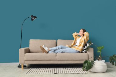 Evdeki mavi duvarın yanındaki gri kanepede yatan genç kadın.