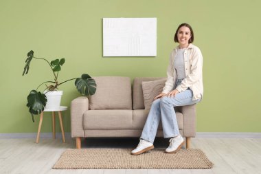 Evdeki yeşil duvarın yanındaki gri koltukta oturan genç kadın.