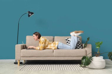 Evdeki mavi duvarın yanındaki gri koltukta dizüstü bilgisayar kullanan genç bir kadın.