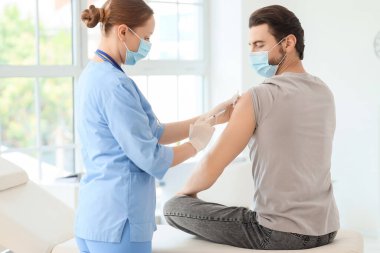 Tıbbi maskeli genç adam klinikteki doktordan aşı alıyor.