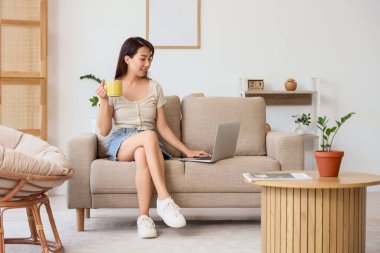 Evdeki kanepede dizüstü bilgisayar kullanan Asyalı genç bir kadın.