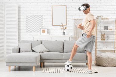 VR gözlüklü genç adam evde futbol topuyla oynuyor.