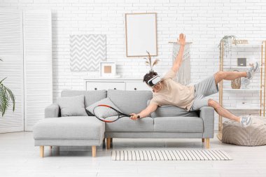 VR gözlüklü genç adam evdeki kanepede tenis oynuyor.