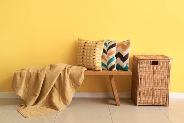 Wooden bench with cushions and wicker basket near yellow wall