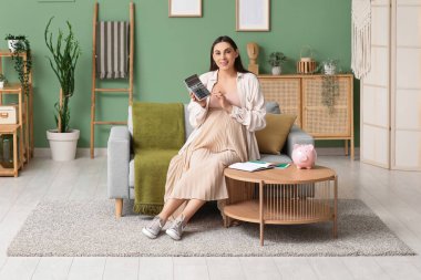 Hesap makinesi, defter ve kumbaralı hamile bir kadın oturma odasındaki kanepede oturuyor.