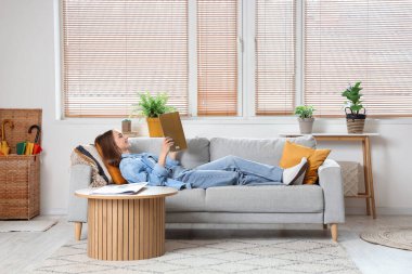 Genç, güzel bir kadın kanepede uzanıyor ve oturma odasında dergi okuyor.