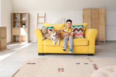 Kulaklıklı şirin Corgi köpeği olan mutlu Asyalı çocuk evdeki kanepede oturuyor.