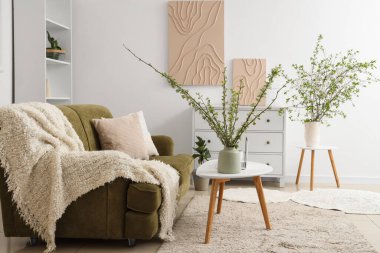 Yeşil kanepe, kahve masası, çekmece sandığı ve aydınlık oturma odasında çiçek açan dalları olan vazo.