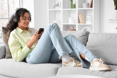 Kulaklıklı genç Afro-Amerikalı kadın evdeki koltukta cep telefonu kullanıyor.