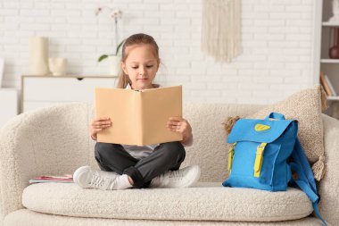 Kitap ve sırt çantasıyla kanepede oturan ve evde ders çalışan sevimli küçük kız.