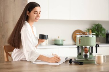 Modern kahve makinesiyle güzel, genç, mutlu bir kadın. Mutfakta kahve içip dergi okuyor.