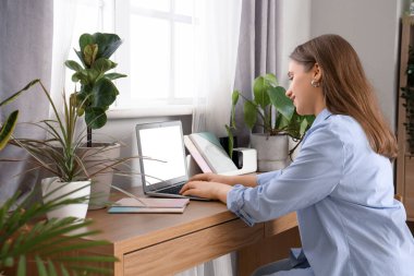 Young woman working with laptop and plants at table near window in office