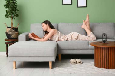 Pijamalı genç kadın kanepede uzanıyor ve oturma odasında kitap okuyor.