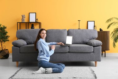 Genç bir kadın kanepenin yanında oturuyor ve oturma odasında dizüstü bilgisayar kullanıyor.