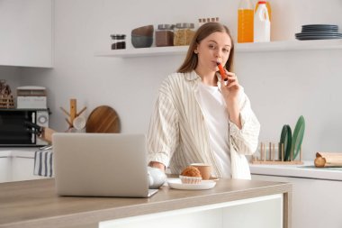 Genç bir kadın mutfakta dizüstü bilgisayar kullanıyor ve elektronik sigara içiyor.
