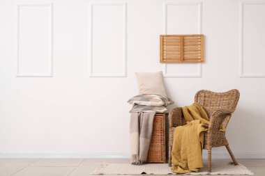 Beyaz duvarın yanında yastık yığını olan hasır koltuk ve sepet.