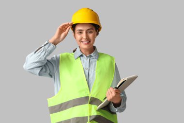 Gri arka planda not defteri ve kalemi olan genç bir inşaat işçisi. Erkek mesleği kavramındaki kadın.