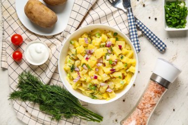 Bir kase lezzetli patates salatası. Beyaz arka planda soğan ve malzemeler var.