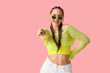 Örgü ören, rastalı ve güneş gözlüklü zarif genç kadın pembe arka plandaki izleyiciyi işaret ediyor.