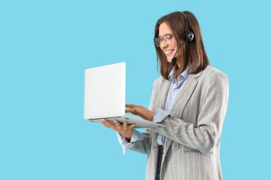 Mavi arka planda dizüstü bilgisayarla çalışan kulaklıklı kadın teknik destek ajanı