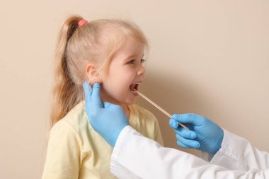 Pediatrician examining little girl with tongue depressor near beige wall