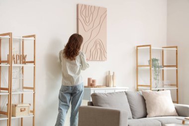 Genç ve güzel bir kadın oturma odasındaki beyaz duvara resim asıyor.