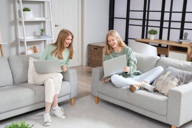 Evdeki kanepede dizüstü bilgisayarı olan güzel, mutlu kız kardeşler.