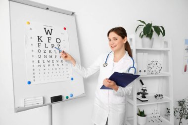 Klinikte göz testi tablosunun yanında panosu olan kadın göz doktoru.