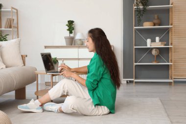 Evdeki sehpada dizüstü bilgisayar kullanan genç bir kadın.