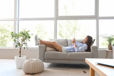 Genç Asyalı kadın evdeki gri koltukta tablet bilgisayar kullanıyor.