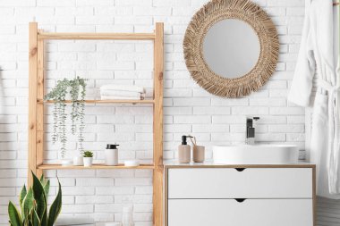 Banyoda beyaz tuğla duvarın yanında havlu ve çiçek bulunan lavabo ve raf ünitesi.