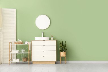 Seramik lavabo, raf ünitesi ve şık banyoda yeşil duvarın yanındaki beyaz çekmeceler.