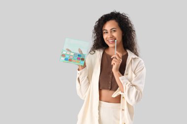 Gri arka planda makyaj fırçası ve göz falı olan güzel, mutlu, Afro-Amerikan kadın.