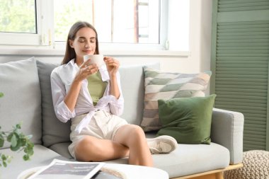 Oturma odasındaki gri koltukta oturan bir fincan kahveyle genç bir kadın.