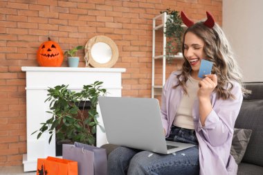 Evde Cadılar Bayramı için internette kredi kartı ve laptop alışverişi yapan şeytan boynuzlu genç bir kadın.