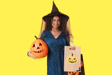 Alışveriş torbaları ve sarı arka planda Cadılar Bayramı balkabağı olan cadı gibi giyinmiş genç bir Afro-Amerikalı kadın.