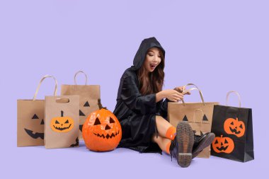 Cadılar Bayramı için giyinmiş genç bir Asyalı kadın, alışveriş torbalarıyla leylak arka planda oturuyor.