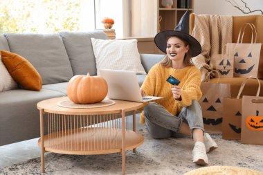 Cadı şapkalı, kredi kartı ve dizüstü bilgisayarı olan genç bir kadın Cadılar Bayramı için internette alışveriş yapıyor.