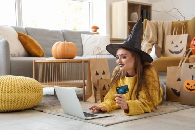 Cadı şapkalı, kredi kartı ve dizüstü bilgisayarı olan genç bir kadın Cadılar Bayramı için internette alışveriş yapıyor.