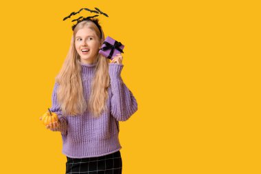 Sarı arka planda balkabağı ve Cadılar Bayramı hediye kutusuyla saç bandı takmış güzel, mutlu genç bir kadın.