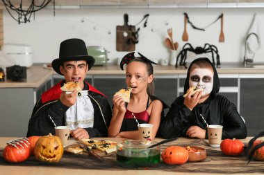 Cadılar Bayramı için giyinmiş çocuklar mutfakta pizza yiyorlar.