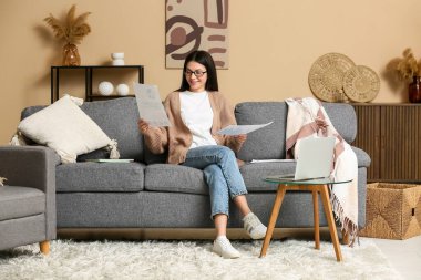 Oturma odasındaki kanepede doküman ve dizüstü bilgisayarla çalışan güzel genç bir kadın.