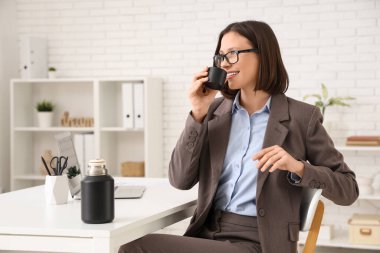 Genç iş kadını çalışma odasında termos bardağından çay içiyor.