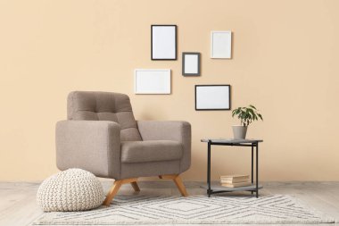 Oturma odasındaki bej duvarın yanında yumuşak puf ve masa olan rahat gri koltuk.