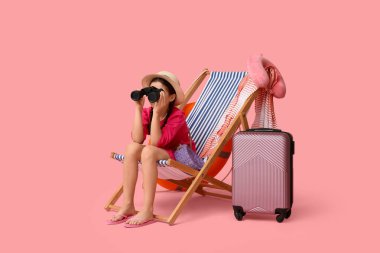 Dürbünü ve bavulu olan sevimli bir kız pembe arka planda iskemlede oturuyor.