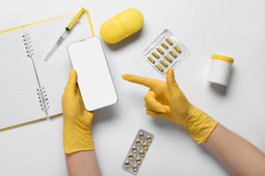 Doktorun elleri gri arka planda defter, hap ve şırıngayla modern cep telefonuna bakıyor.