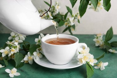 Çaydanlıktan fincana sıcak yasemin çayı ve yeşil masada çiçekler dökülüyor.