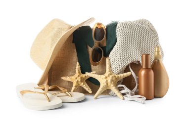 Hasır şapkalı plaj çantası, deniz yıldızları, parmak arası terlikler ve beyaz arka planda güneş kremi şişeleri.